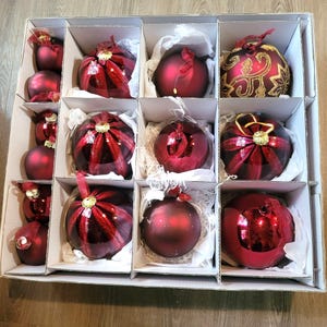Puede incluir: Una colección de adornos navideños rojos variados almacenados en una caja de cartón compartimentada. El conjunto incluye esferas rojas mate, brillantes y esferas rojas con detalles dorados o rojos brillantes, cada una con una tapa dorada para colgar.