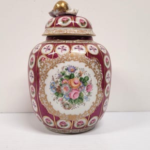 May include: A porcelain ginger jar with a lid featuring a gold finial. The jar has a deep magenta background with white circular medallions containing floral motifs. A central white panel displays a colorful bouquet of roses and daisies.