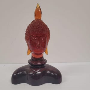 May include: A reddish-brown Buddha head statue with a pointed top and a detailed face. The statue has a dark brown base and a light brown halo. The statue is made of glass or a similar material.