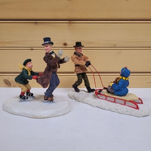 Puede incluir: Un conjunto de tres figuras con temática invernal. Un hombre con un sombrero de copa y un abrigo marrón está patinando sobre hielo con un niño con una chaqueta verde. Otro hombre con una chaqueta marrón está tirando de un trineo con un niño con un abrigo azul.