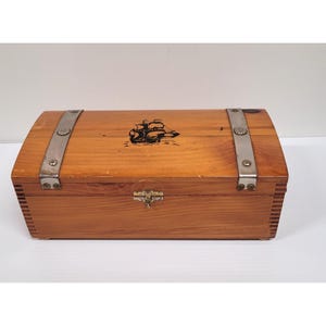 May include: A wooden chest with a curved top, featuring a black ship illustration. The chest has metal bands and a clasp closure. The wood has a warm, natural tone.