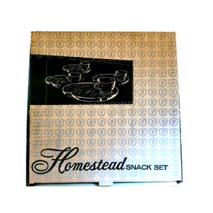 May include: A vintage Homestead Snack Set box. The box has a repeating pattern of silver crests on a light background. A black panel displays a graphic of teacups and trays. The words "Homestead SNACK SET" are printed on the box.