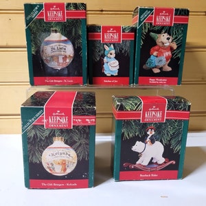 May include: Five Hallmark Keepsake Ornament boxes with different Christmas themes. The boxes are red and green with gold lettering. The ornaments are visible through the clear plastic windows on the front of the boxes. The ornaments include a glass ball with a St. Lucia scene, a glass ball with a Kolyada scene, a porcelain figurine of a bunny, a porcelain figurine of a bear riding a polar bear, and a porcelain figurine of a woodcutter.