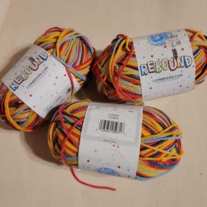 May include: Three skeins of Lion Brand Rebound yarn in the Tigger colorway. The yarn is a mix of yellow, red, blue, and black. Each skein is labeled with the Lion Brand logo and product information.
