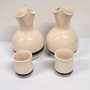 May include: A set of four ceramic pieces: two cream-colored, bulbous vases with ruffled rims and two matching small cups. Each piece has a dark brown band around the base, creating a cohesive design. The set is likely for decorative or functional use.