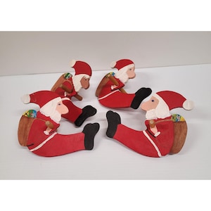 Puede incluir: Cuatro figuras de madera de Papá Noel, cada una en posición sentada con un traje rojo, botas negras y barba blanca. Cada Papá Noel tiene un saco marrón lleno de regalos coloridos y un gorro rojo. Las figuras están pintadas con un acabado rústico.