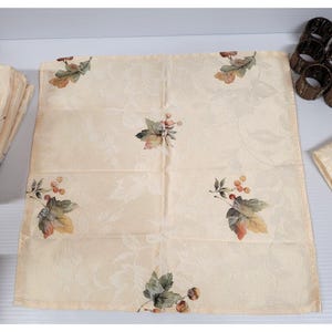 May include: Cream-colored cloth napkin with a floral and leaf pattern. The napkin features a repeating design of leaves and berries in shades of green, orange, and brown. A set of matching napkin rings is visible.
