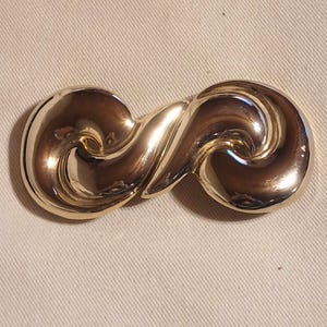 Puede incluir: Un broche de metal dorado con un diseño abstracto y ondulado. El broche pulido tiene una forma simétrica, con dos extremos redondeados conectados por un centro curvo. El diseño recuerda a un símbolo de infinito.