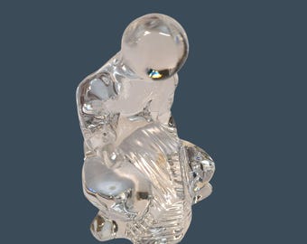 May include: A clear glass sculpture of a person in a kneeling position. The figure has a rounded head and is bent over, with the arms and legs in a folded position. The sculpture is transparent and reflects light.