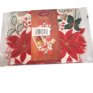 Könnte beinhalten: Ein St. Nicholas Square Tischläufer in einer transparenten Kunststoffverpackung. Der Läufer zeigt rote Weihnachtssterne, grüne Stechpalmenblätter und Tannenzapfen auf weißem Grund. Die Maße betragen 91,4 cm mal 33 cm.