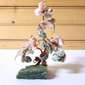 May include: A small decorative tree made of seashells and wire. The tree has pink flowers and green leaves. The tree is sitting on a green stone base.