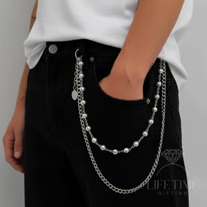 May include: Silver-toned chain with pearl and chain accents, attached to black denim jeans. The chain features multiple strands of small pearls and a longer chain, creating a layered effect. A small, round charm hangs from the top.