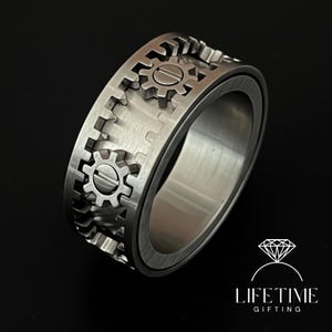以下が含まれることがあります： 歯車デザインのシルバーカラーのリング。リングは、バンドの周りに複数の小さな歯車の形をしており、一部の中央にはネジのディテールがあります。リングはつや消しメタル仕上げで、背景は暗いです。