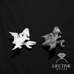 May include: Two silver dragon rings. One ring is a solid silver color, and the other has a darker, textured appearance with red accents. The rings are set against a black background.