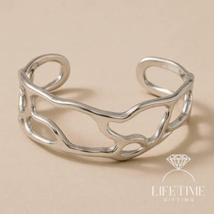 May include: A silver-colored cuff bracelet with an open, organic design. The bracelet features irregular, flowing shapes and a polished finish. The open design allows for easy wear. The bracelet is a piece of jewelry.