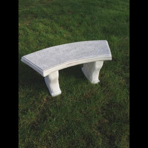May include: A curved, light grey concrete garden bench with a textured surface. The bench has two decorative legs and is set on green grass. The bench is approximately 122 cm long.