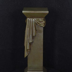 Op de afbeelding: Een bronzen sokkel met een vierkante boven- en onderkant, met een gedrapeerd stoffen ontwerp en Grieks sleutelpatroon. De sokkel is een decoratief item, waarschijnlijk voor het tentoonstellen van sculpturen of andere objecten.
