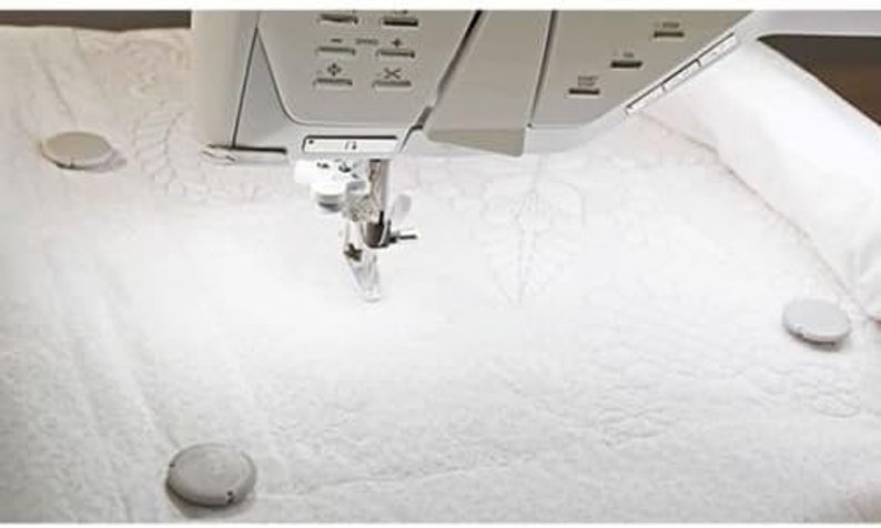 Puede incluir: Una m&aacute;quina de coser blanca con una aguja e hilo en su lugar, lista para coser. La m&aacute;quina est&aacute; sobre una superficie blanca con tres objetos redondos blancos.