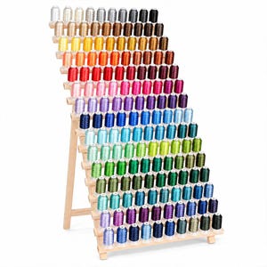 May include: A tiered wooden display rack holding a large collection of sewing thread spools arranged in a gradient of colors. The spools are organized in rows, featuring a wide spectrum including white, gray, black, yellow, orange, red, pink, purple, blue, and green shades.