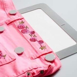 Puede incluir: Tela vaquera rosa con dise&ntilde;o floral bordado y botones blancos. La tela est&aacute; sujeta por un aro de bordado gris.