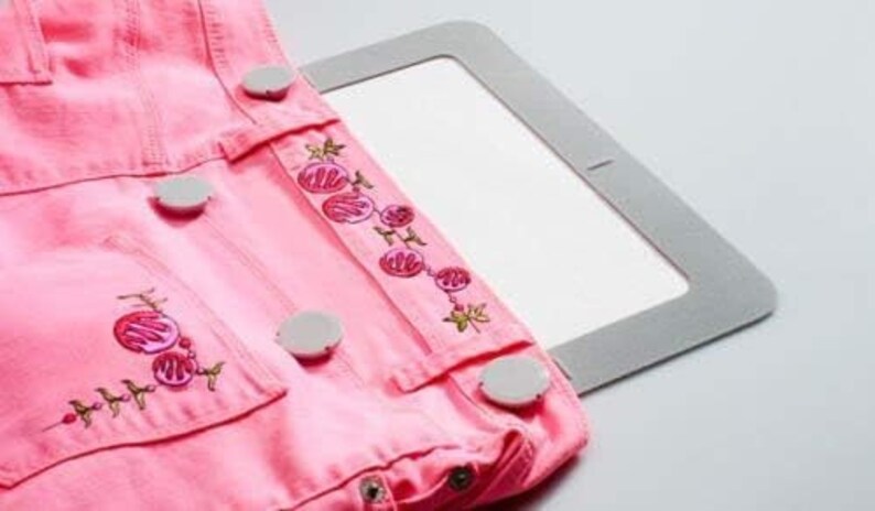 Puede incluir: Tela vaquera rosa con dise&ntilde;o floral bordado y botones blancos. La tela est&aacute; sujeta por un aro de bordado gris.