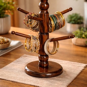 May include: A dark brown wooden jewelry stand with multiple horizontal pegs for displaying bracelets. The stand holds various bracelets in gold, silver, and colorful designs. The stand is on a beige placemat on a wooden table.