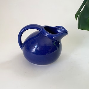 May include: A small, cobalt blue ceramic creamer or pitcher with a rounded body and a curved spout. The handle is attached to the side of the pitcher.