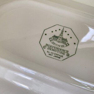 May include: Close-up of a white ceramic dish with a green logo that reads "Independence Ironstone" with a copyright symbol, "1776", a registered trademark symbol, and "by Interpace NKT Japan".