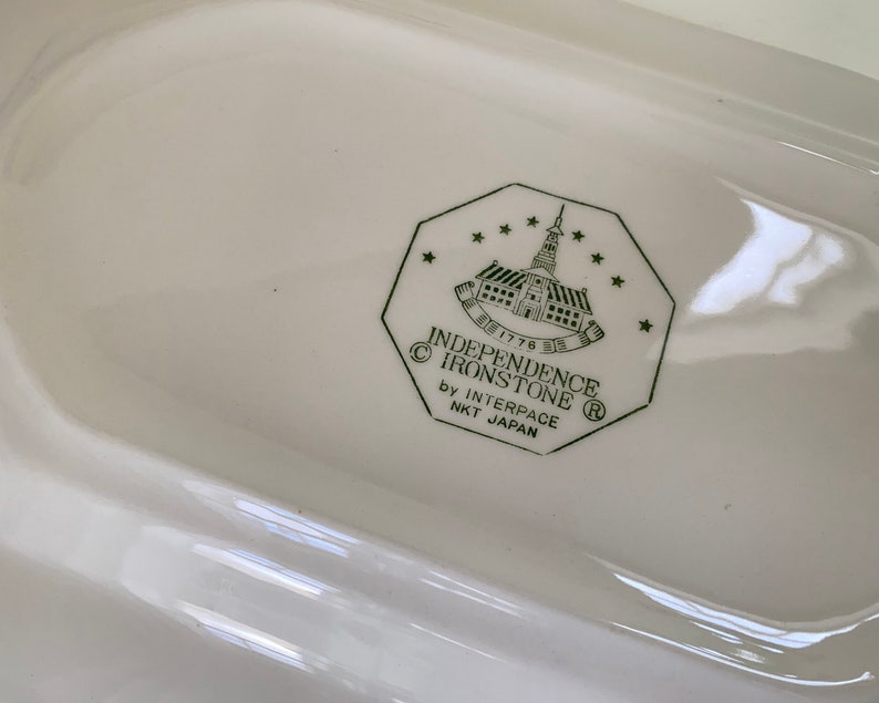 May include: Close-up of a white ceramic dish with a green logo that reads "Independence Ironstone" with a copyright symbol, "1776", a registered trademark symbol, and "by Interpace NKT Japan".
