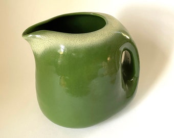 May include: A green ceramic pitcher with a white rim. The pitcher has a unique, rounded shape and a handle.