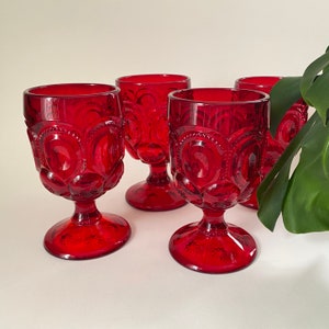 May include: Four ruby red vintage wine glasses. Each glass features a detailed pattern of circles and crescent shapes. The stemware has a footed base with a starburst design. The glasses are arranged on a white surface.