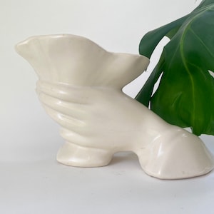 May include: White ceramic hand-shaped vase with a ruffled opening. The vase is a modern design and would be a great addition to any home decor.