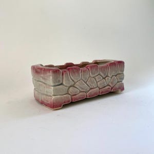 May include: A rectangular ceramic planter with a stone-like texture. The planter features a light gray base with pink accents on the edges and rim. The design resembles stacked stones, suitable for small plants or succulents.