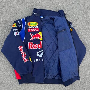 Op de afbeelding: Marineblauwe jas met diverse sponsorlogo's, waaronder Red Bull, Geox en Infiniti. De jas heeft een gewatteerde binnenkant en een opstaande kraag. De mouwen tonen het Red Bull logo.