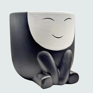 May include: A black and white ceramic planter with a smiling face design. The planter has a rounded body with a white face and black accents, including the eyes and legs. The planter is designed to look like a character sitting down.