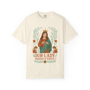 May include: Cream-colored t-shirt featuring a graphic of Our Lady undoing knots, with angels and floral accents. The design includes the text "OUR LADY UNDOER OF KNOTS" in a decorative font. The shirt has short sleeves and a crew neck.