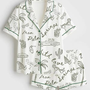 May include: White pajama set with a green trim and a Palm Springs-themed print. The print includes illustrations of cocktails, flamingos, palm trees, and the text "With Love From Palm Springs 2026". The set includes a button-down shirt and shorts.
