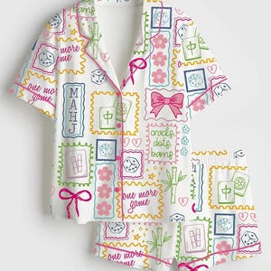 May include: A white short-sleeve button-down pajama shirt and matching shorts set featuring a colorful print of mahjong tiles, dice, pink cherry blossoms, bows, bamboo stalks, and the text "one more game" and "cracks dots bams" in script.