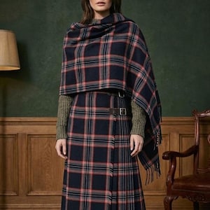 Può includere: Una donna indossa uno scialle e una gonna abbinata a quadri blu navy, rossi e bianchi. La gonna è cinta in vita con una cintura in pelle marrone. L'outfit è abbinato a un maglione verde. Lo scialle ha un bordo sfrangiato.