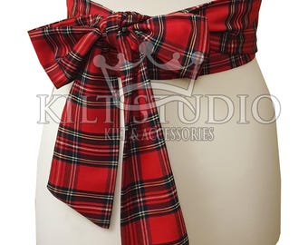 May include: A red tartan sash with a large bow, displayed on a mannequin. The sash features a classic plaid pattern in red, black, and white. The text "Kilt Studio" is visible.