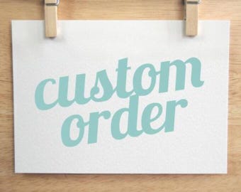 May include: A rectangular white card featuring the text custom order printed in a light teal sans-serif font. The card is held by two wooden clips at the top edge, displaying the words clearly against the plain white background surface.