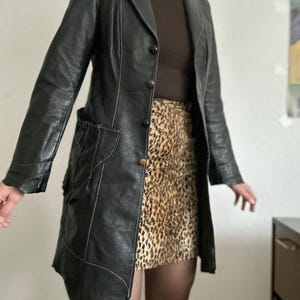May include: Black leather coat with white stitching details, featuring a notched collar and button closure. The coat is paired with a brown turtleneck and a leopard print skirt. The coat has a unique asymmetrical hemline.