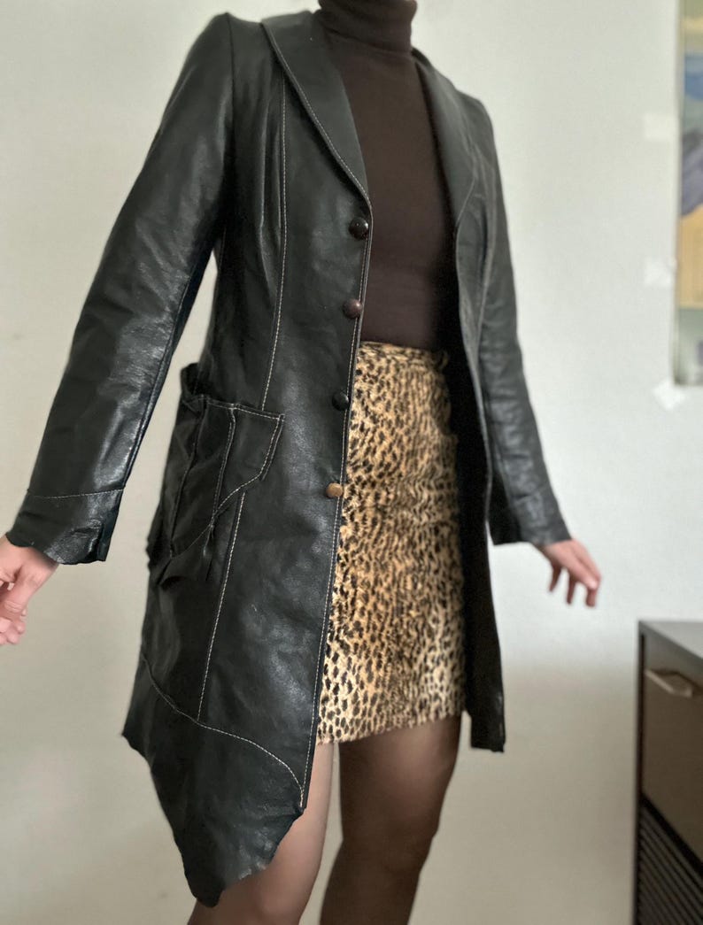 May include: Black leather coat with white stitching details, featuring a notched collar and button closure. The coat is paired with a brown turtleneck and a leopard print skirt. The coat has a unique asymmetrical hemline.
