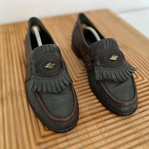 Puede incluir: Un par de mocasines de cuero gris oscuro con ribetes y flecos marrones. Cada zapato presenta un emblema decorativo y una correa de cuero. Los zapatos se exhiben en una tabla de cortar de madera.