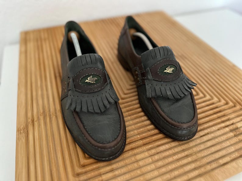 Puede incluir: Un par de mocasines de cuero gris oscuro con ribetes y flecos marrones. Cada zapato presenta un emblema decorativo y una correa de cuero. Los zapatos se exhiben en una tabla de cortar de madera.