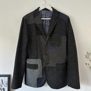 May include: A dark gray blazer with a patchwork design. The blazer features a mix of textures and patterns, including houndstooth and solid gray panels. It has a two-button closure and a notched lapel.