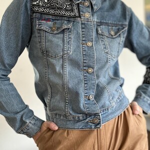 Op de afbeelding: Lichtblauwe spijkerjas met een zwart-wit paisley patroon op de kraag en schouders. De jas heeft knoopsluitingen, twee borstzakken en lange mouwen. De jas wordt gedragen met een bruine broek.