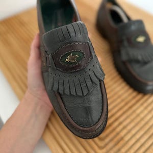 Puede incluir: Mocasines de cuero verde oscuro con ribetes y flecos marrones. Los zapatos tienen un emblema decorativo con un p&aacute;jaro dorado. Los zapatos se exhiben sobre una superficie de madera.
