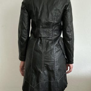 May include: Black leather coat with white stitching details. The coat features a notched collar, long sleeves, and a belted waist. The coat has a flared skirt and is knee-length.