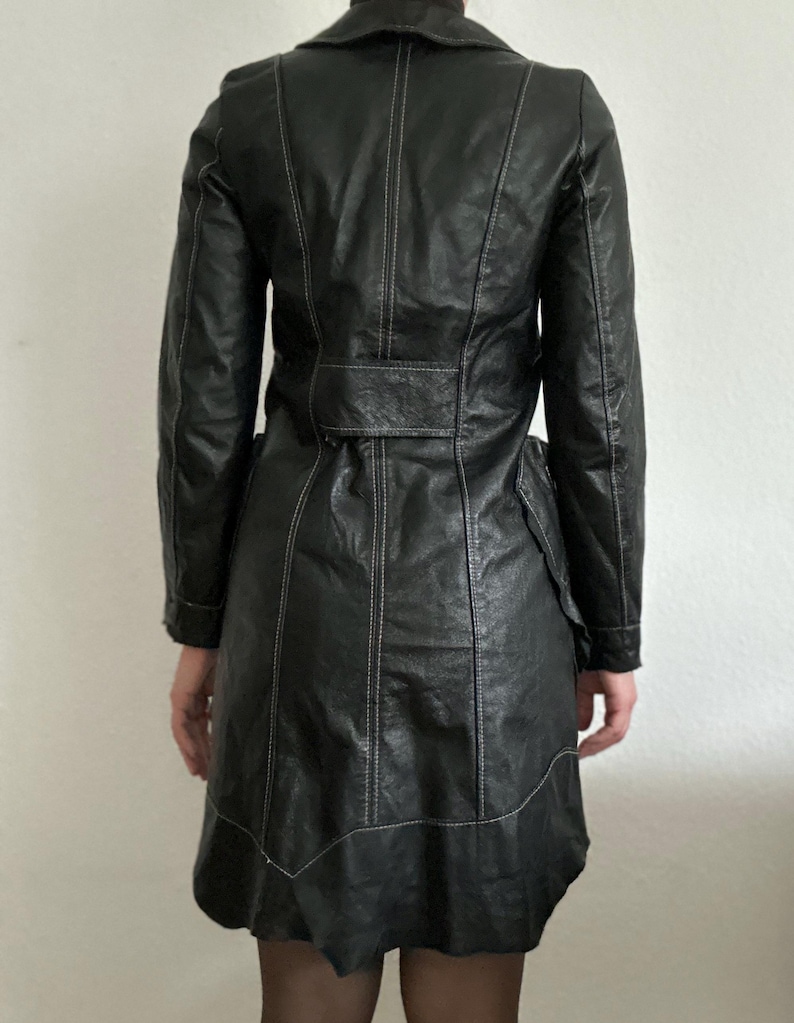 May include: Black leather coat with white stitching details. The coat features a notched collar, long sleeves, and a belted waist. The coat has a flared skirt and is knee-length.